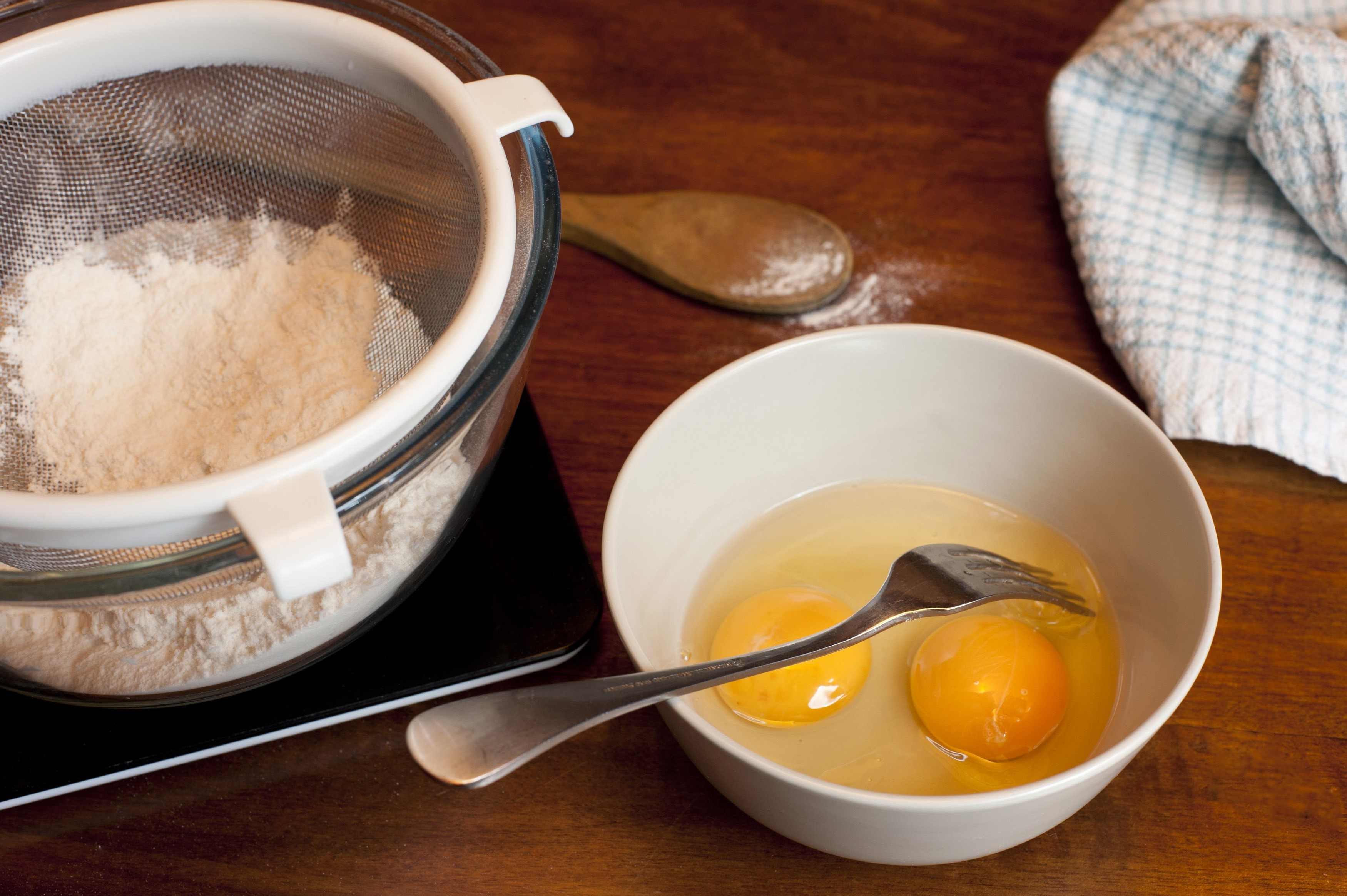 Fresh baking ingredients with two eggs in a mixing bowl and sifted white cake flour in a sieve on a wooden kitchen counter